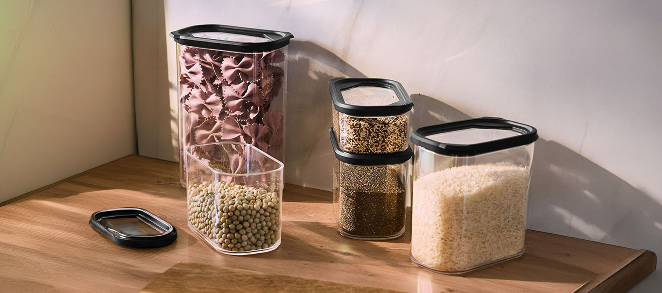 Recipientes de cozinha transparentes com tampas pretas, cheios de massa, lentilhas, quinoa, sementes de chia e arroz numa superfície de madeira.