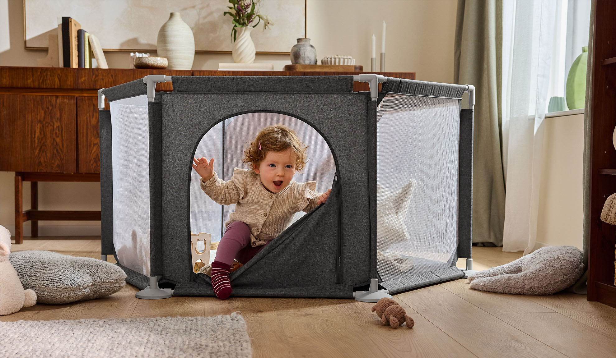 Bebé feliz a sair de um parque cinzento numa sala de estar acolhedora.