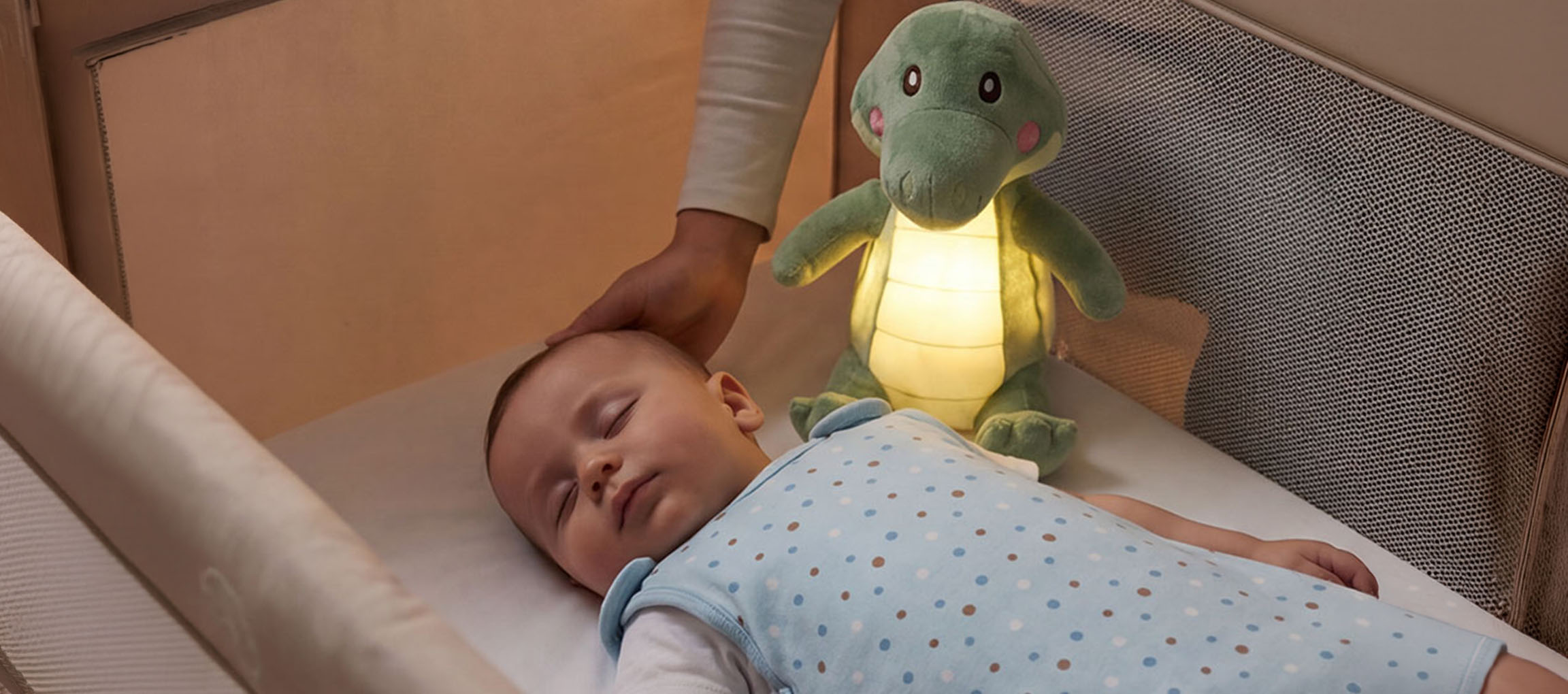 Bebé a dormir num berço com um peluche luminoso, adulto a acariciar a cabeça do bebé.