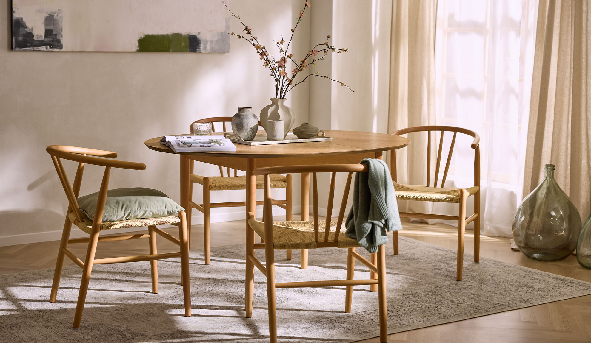 Sala de jantar luminosa com mesa redonda de madeira, cadeiras de madeira com assentos de corda e decoração floral.