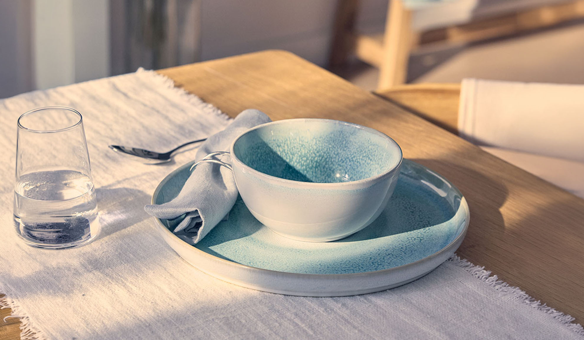 Mesa posta com uma tigela azul, prato, copo de água e guardanapo numa mesa de madeira.
