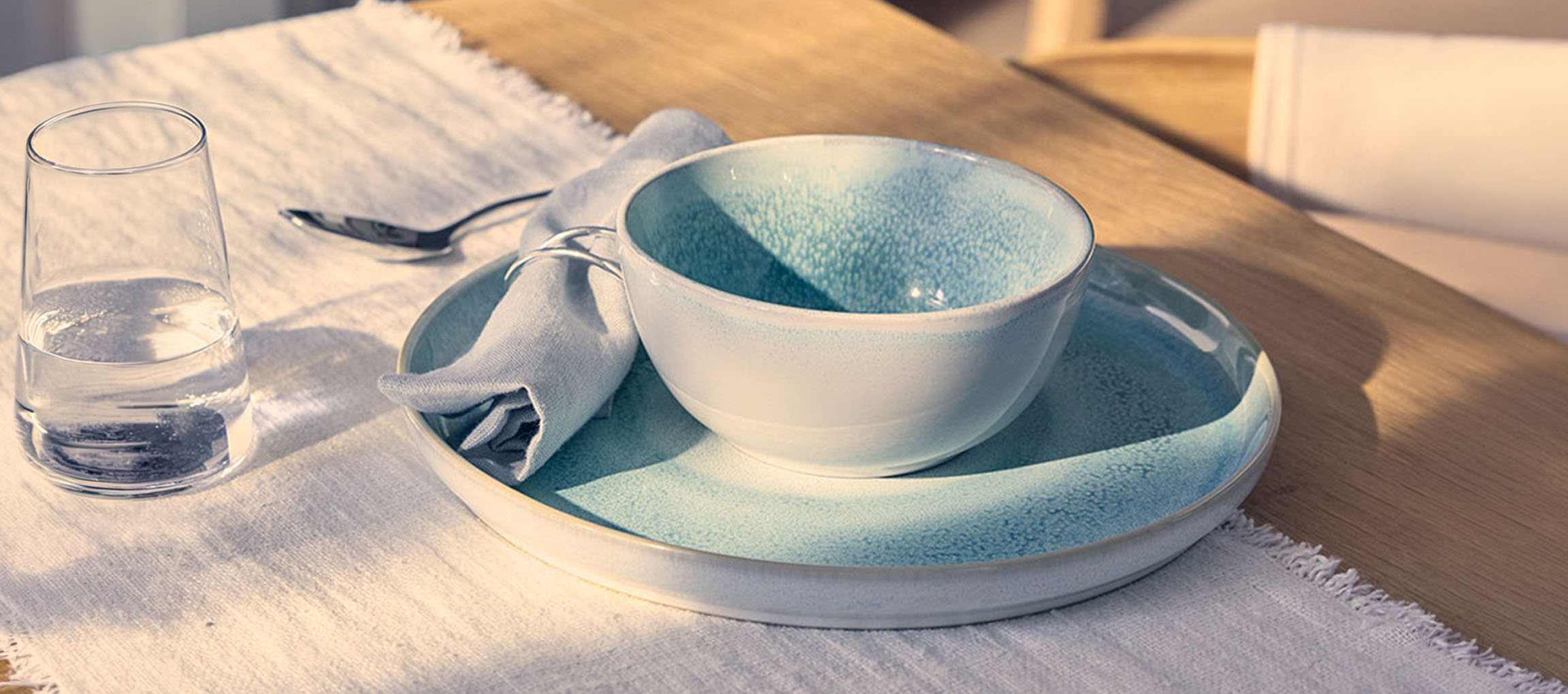 Mesa posta com uma tigela e prato de cerâmica azul, copo de água e guardanapo.