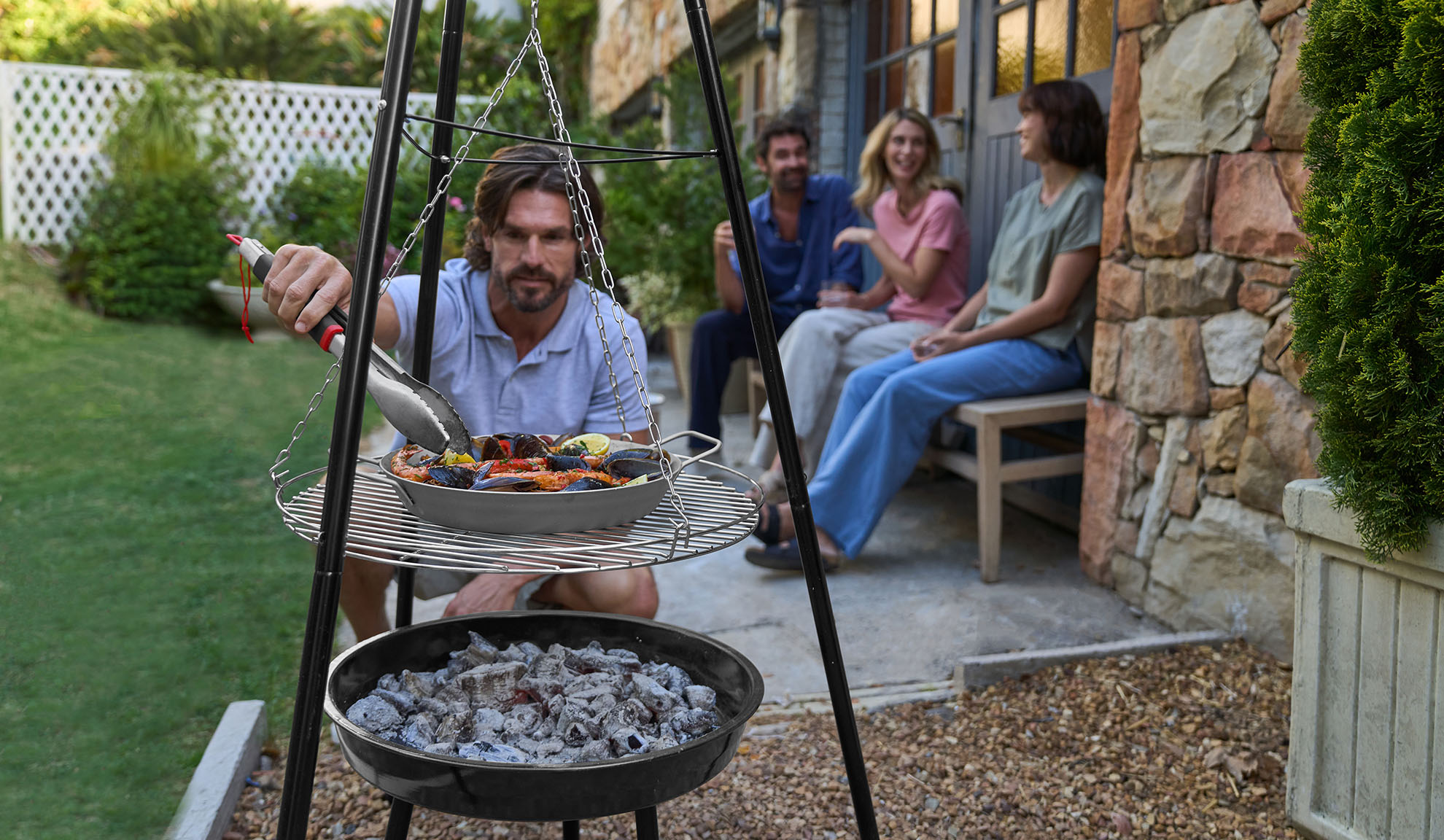 Homem grelhando marisco num tripé, amigos a relaxar ao fundo.