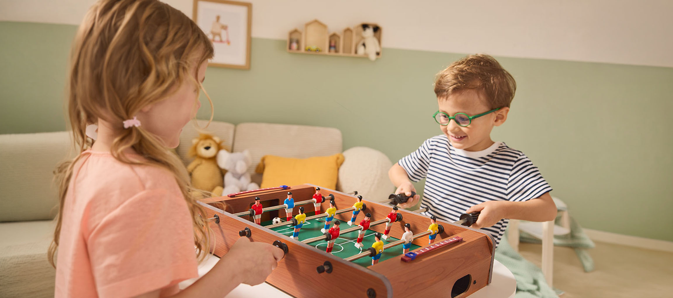 Duas crianças a jogar matraquilhos, uma menina de camisola rosa e um menino de camisola às riscas.
