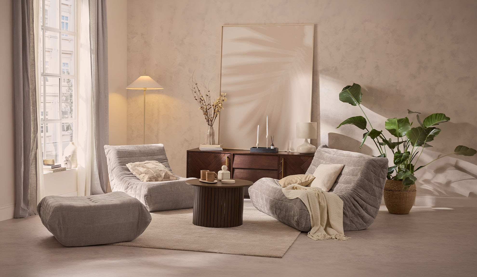 Sala de estar acolhedora com duas cadeiras de lounge cinzentas, um pufe, uma mesa de centro e uma planta.