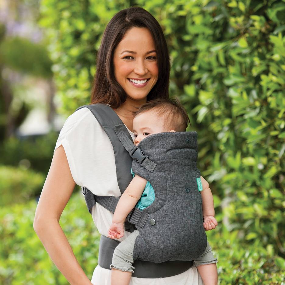 Mulher sorridente com um porta-bebés cinzento a segurar um bebé ao ar livre.