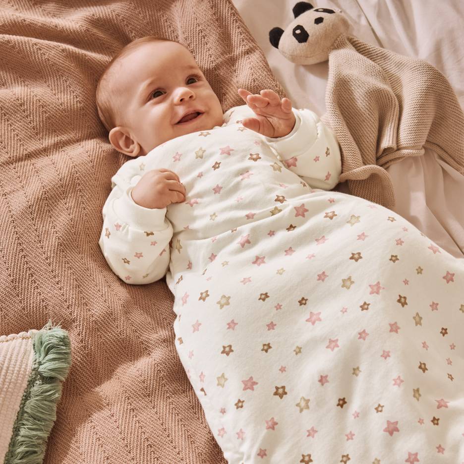 Bebé sorridente em saco de dormir branco com estrelas rosa e castanhas, deitado numa manta.