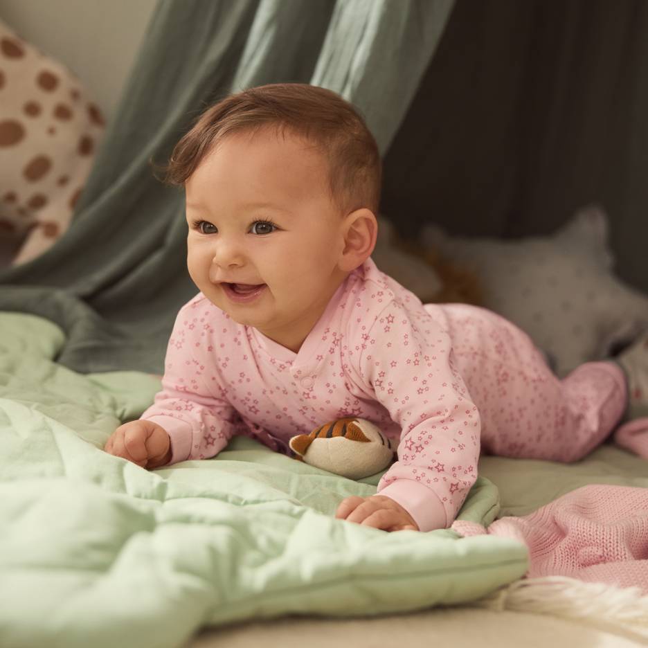 Bebé de pijama rosa com estrelas, deitado numa manta verde com um peluche.
