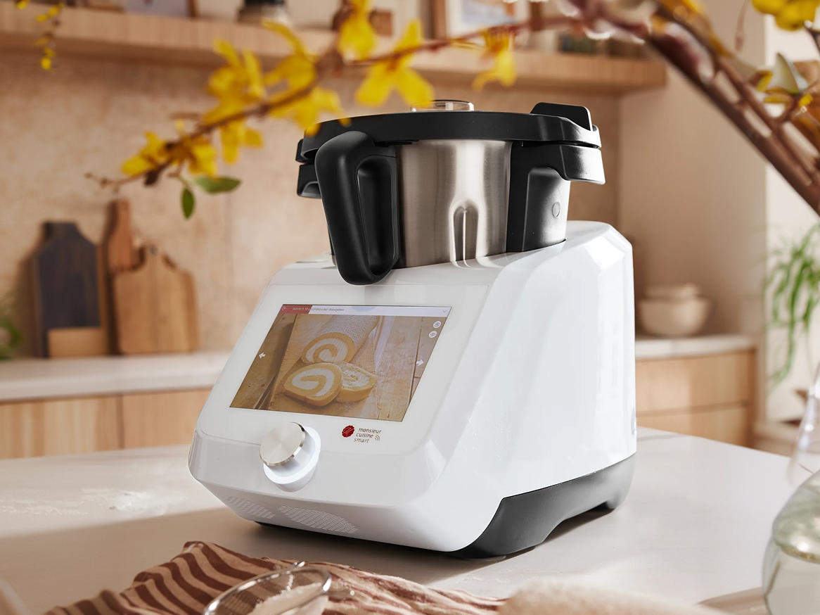 Robô de cozinha branco com ecrã tátil a exibir um bolo suíço, em bancada de cozinha.