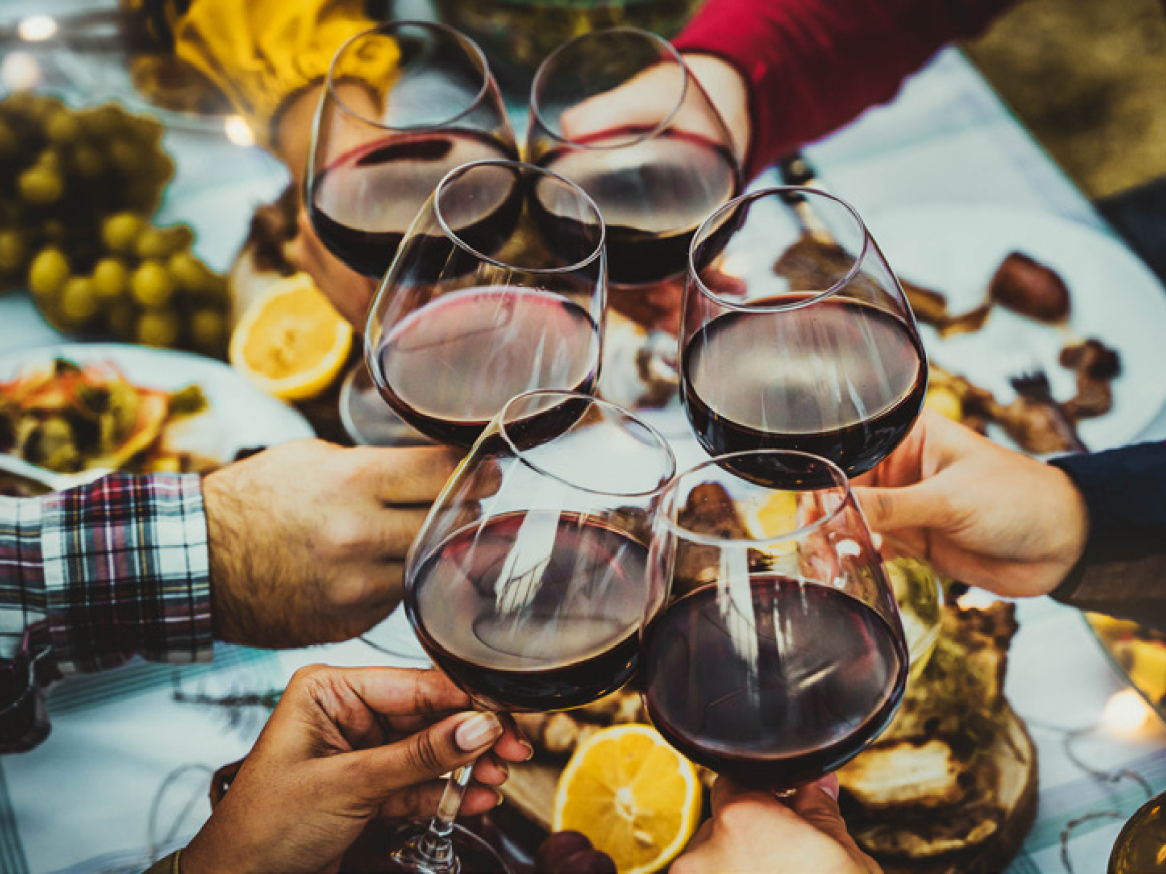 Pessoas a brindar com copos de vinho tinto numa mesa de jantar com comida.