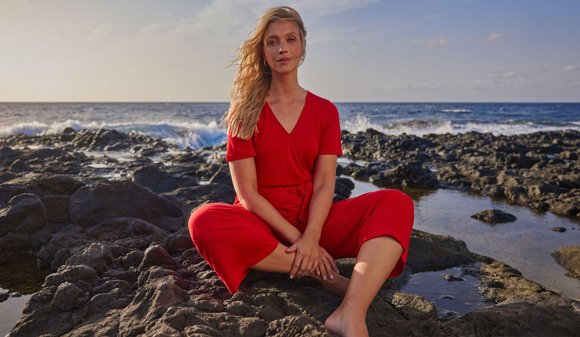 Mulher de macacão vermelho sentada em rochas junto ao mar.