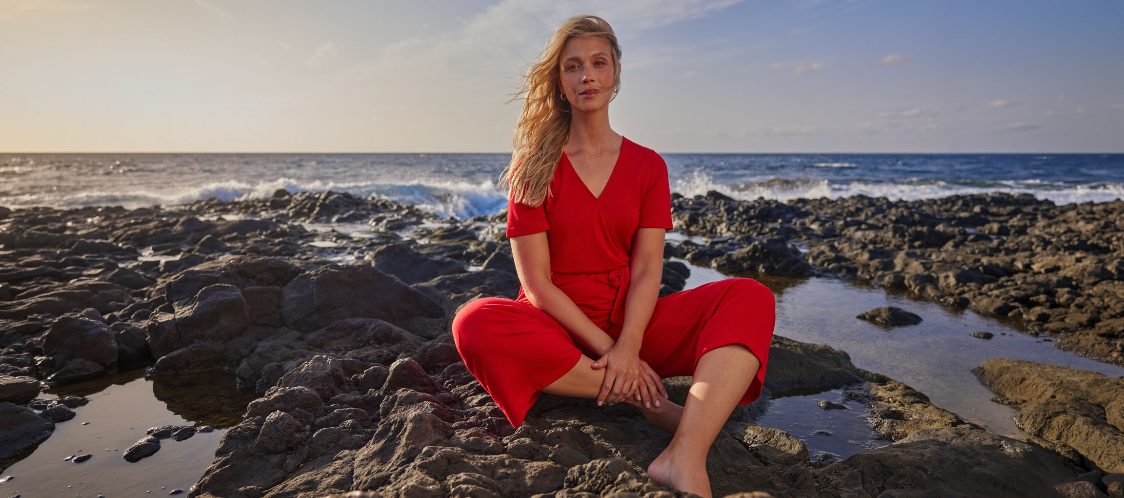 Mulher de macacão vermelho sentada numa praia rochosa junto ao mar.