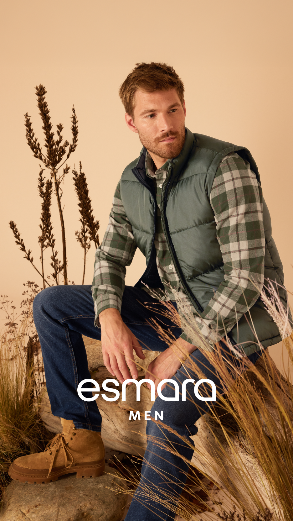 Homem com colete verde, camisa xadrez, jeans e botas, sentado na natureza.