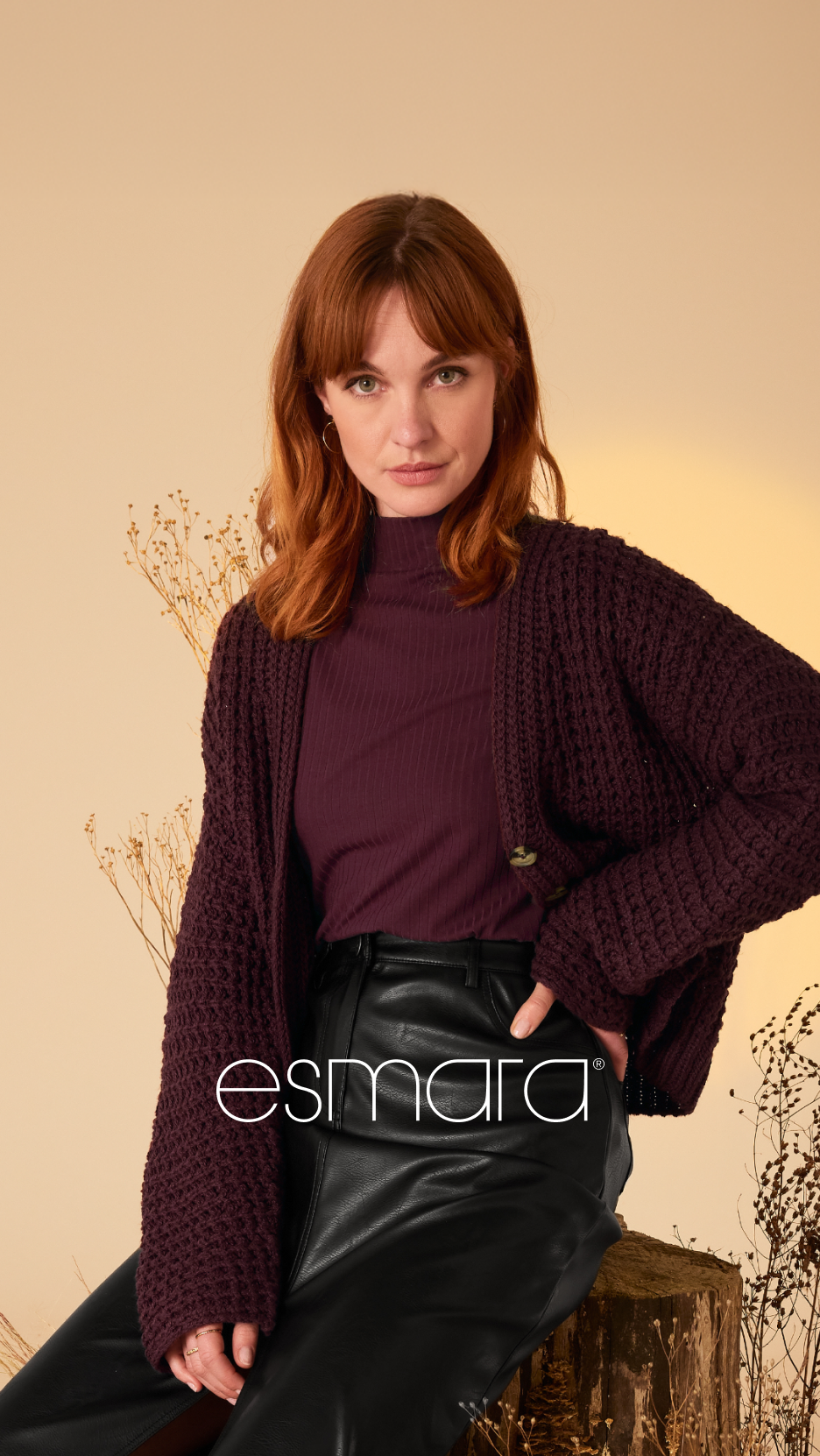 Uma mulher de cabelo ruivo a usar um casaco de malha roxo, uma camisola de gola alta e calças de couro pretas.
