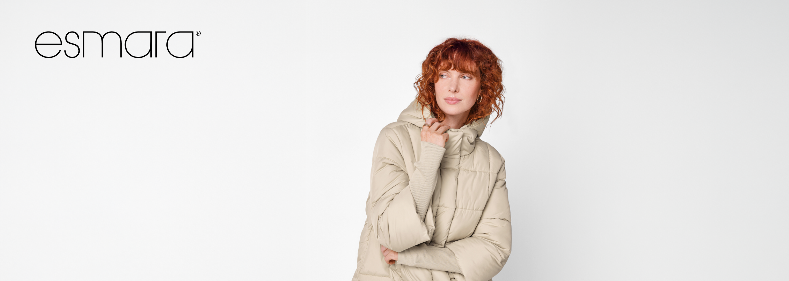 Mulher com cabelo ruivo encaracolado, casaco acolchoado bege, logótipo Esmara.