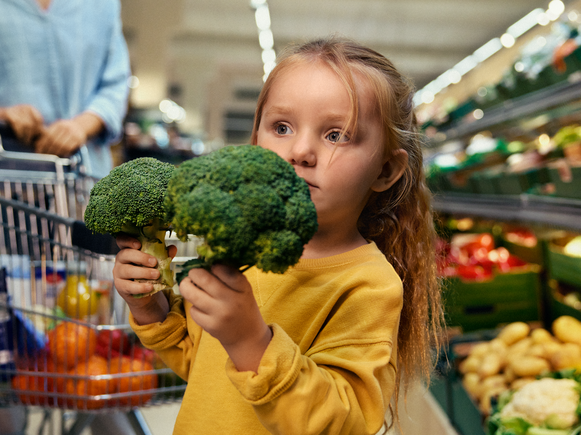 Criança a segurar brócolos frescos num supermercado, com um carrinho de compras ao fundo.