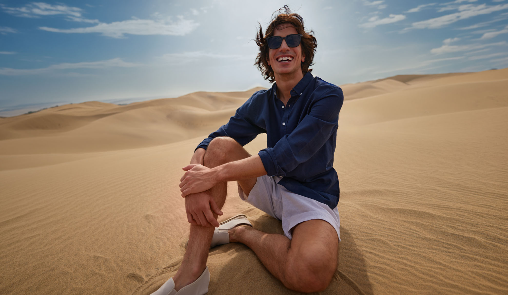 Homem de camisa azul escura e calções claros sentado nas dunas de areia, a sorrir.