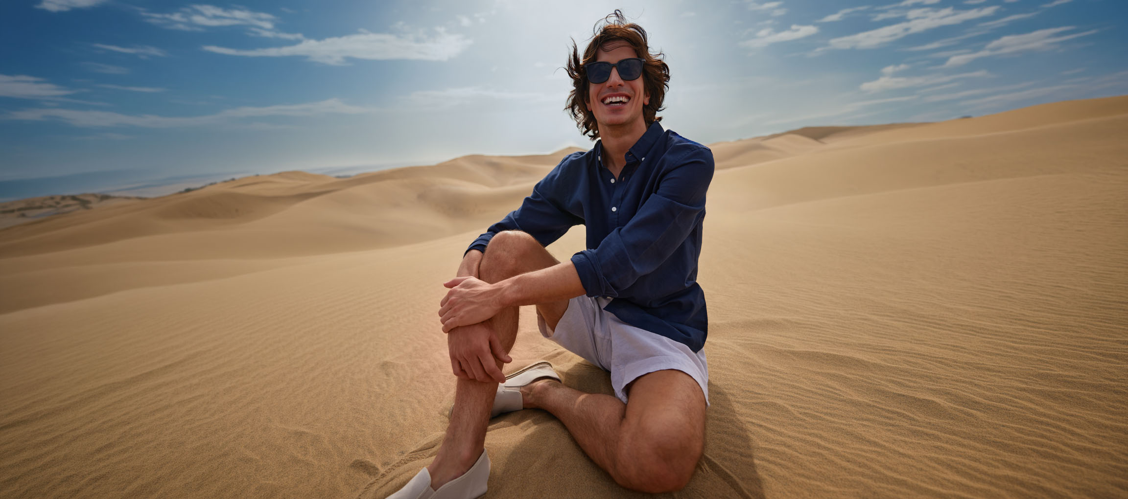 Homem de camisa azul escura e calções sentado nas dunas de areia, a sorrir.