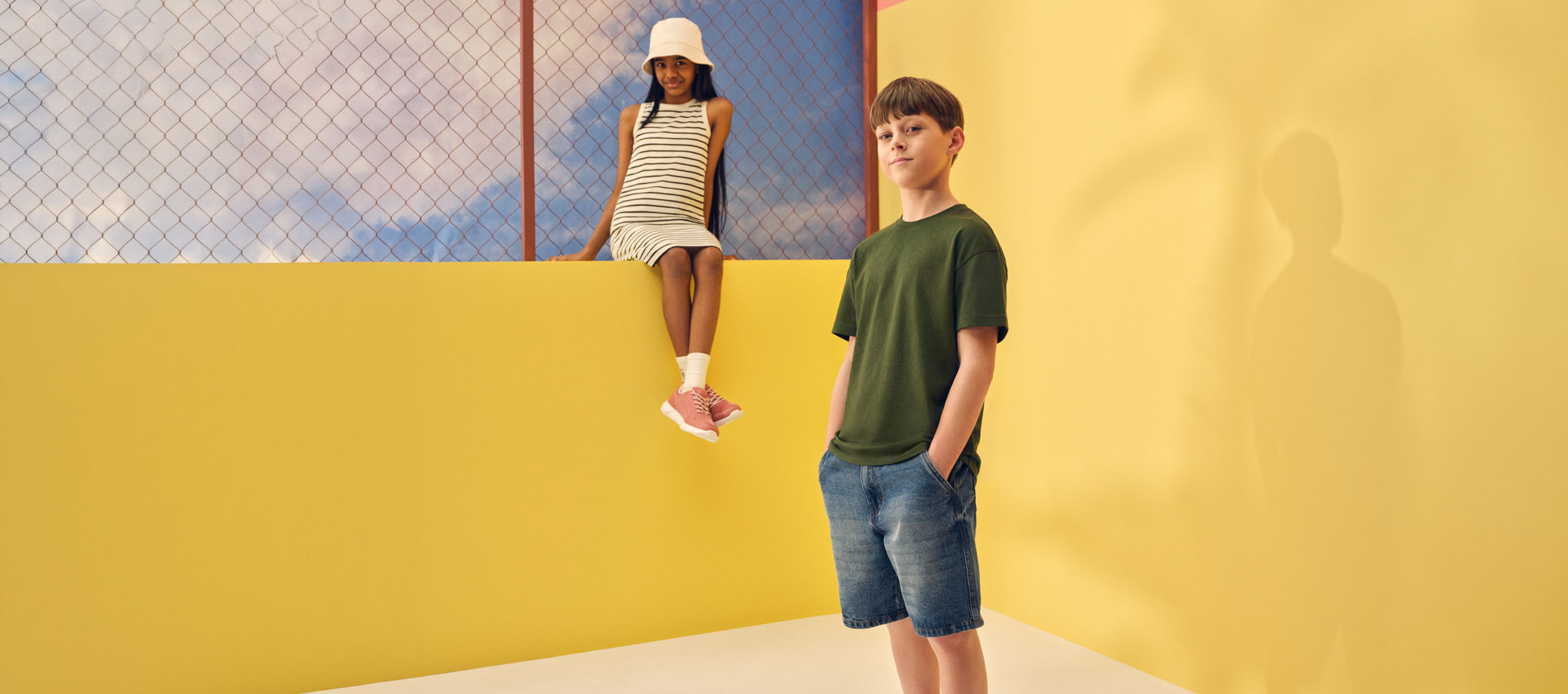 Duas crianças com roupas casuais de verão: uma menina com um vestido às riscas e um menino com uma t-shirt verde e calções de ganga.