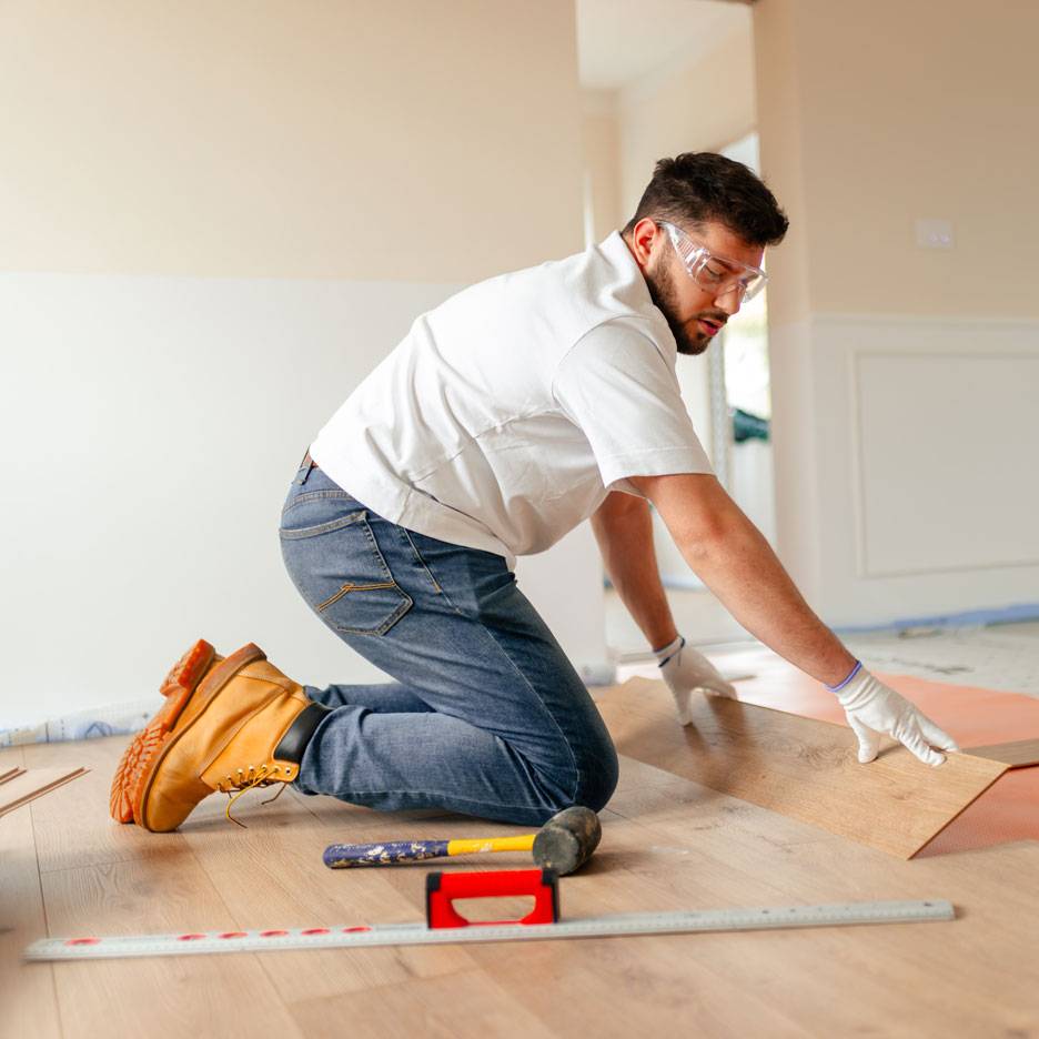Homem a instalar pavimento laminado, usando óculos de segurança e botas de trabalho.