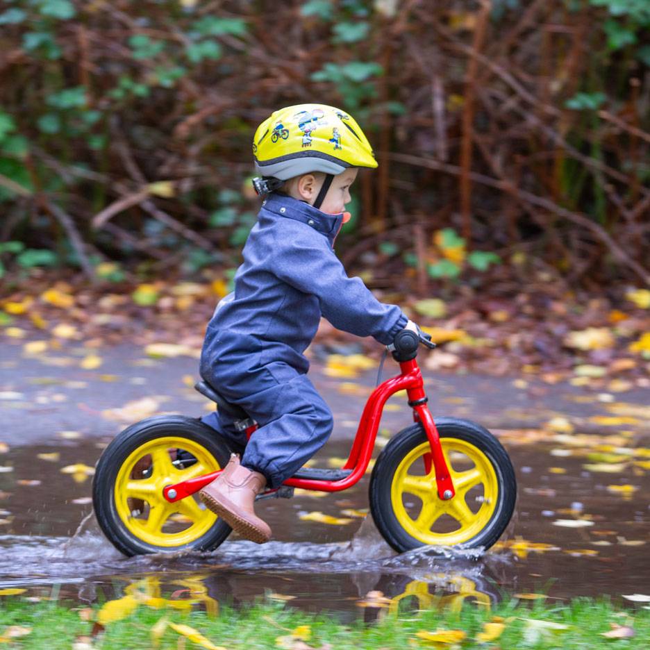 Criança em bicicleta de equilíbrio numa poça, com capacete e macacão.