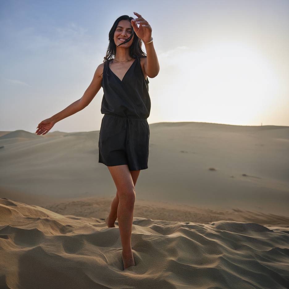Mulher de macacão preto descalça em dunas de areia ao pôr do sol.