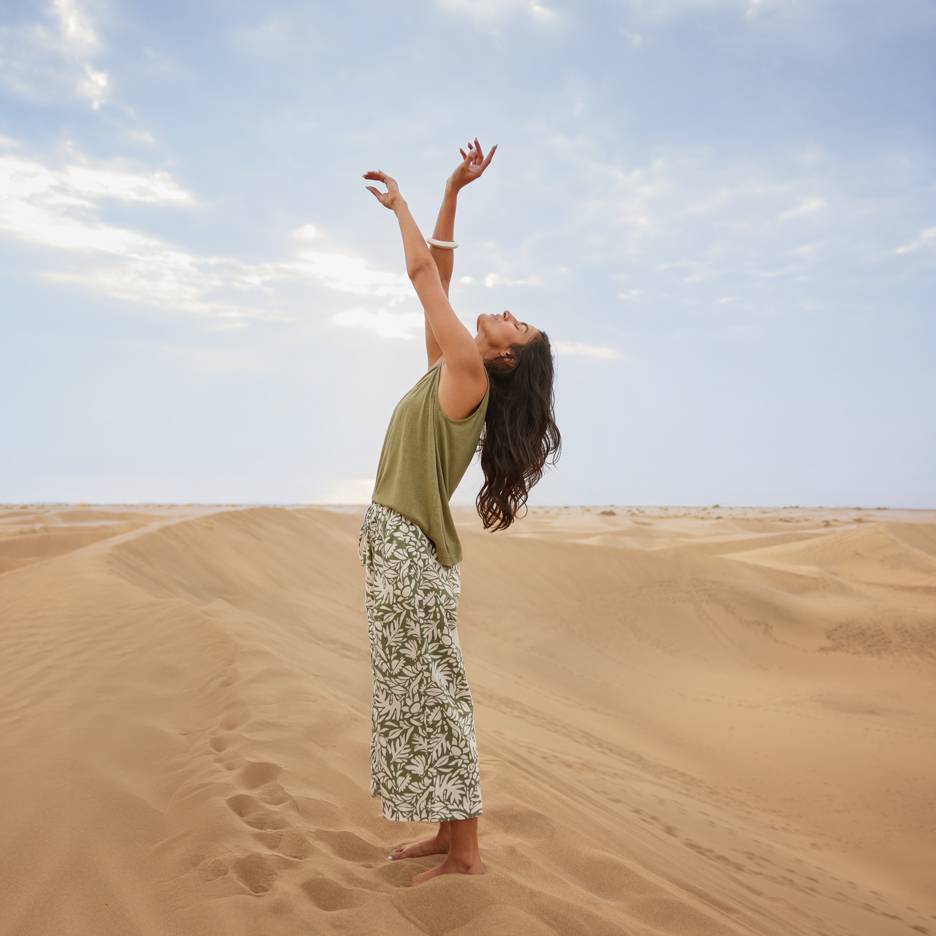 Mulher nas dunas de areia com top verde e calças estampadas, braços levantados.