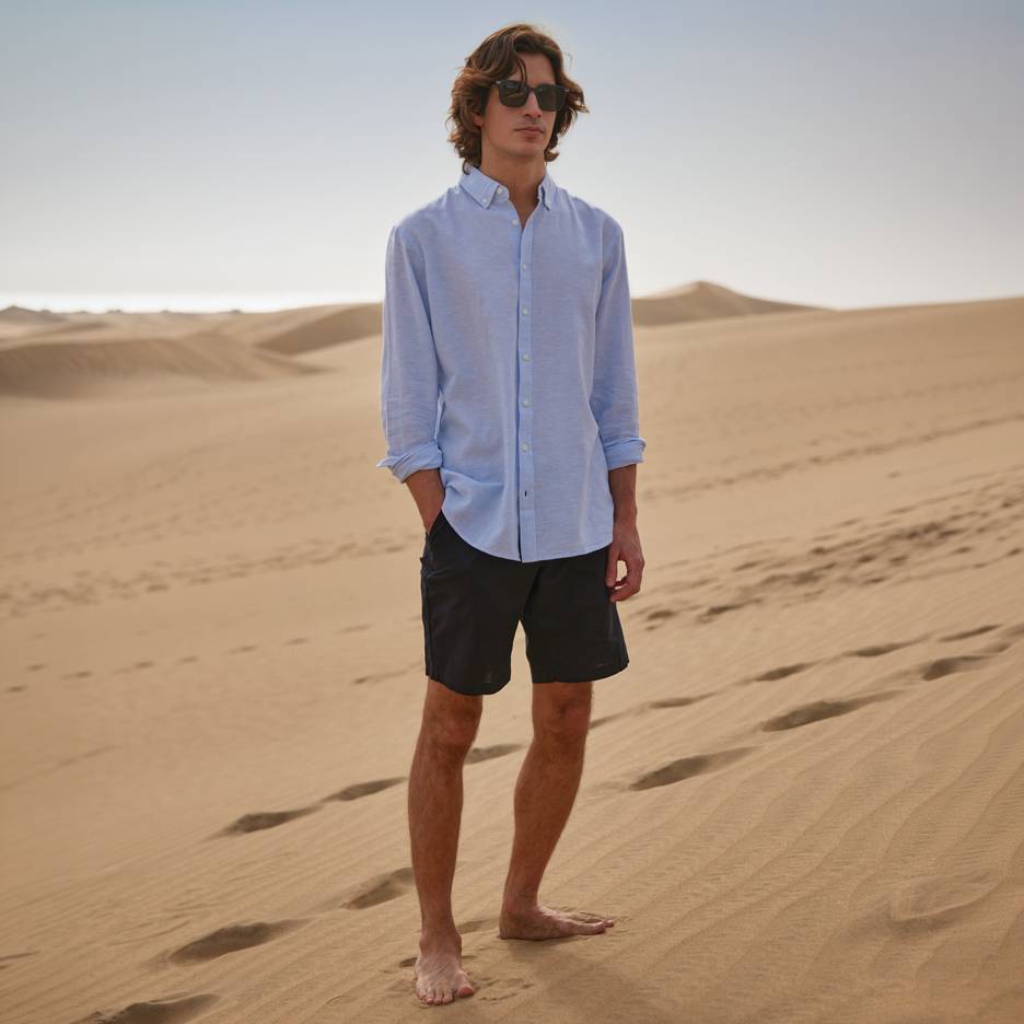 Homem de camisa azul e calções pretos, descalço em dunas de areia.
