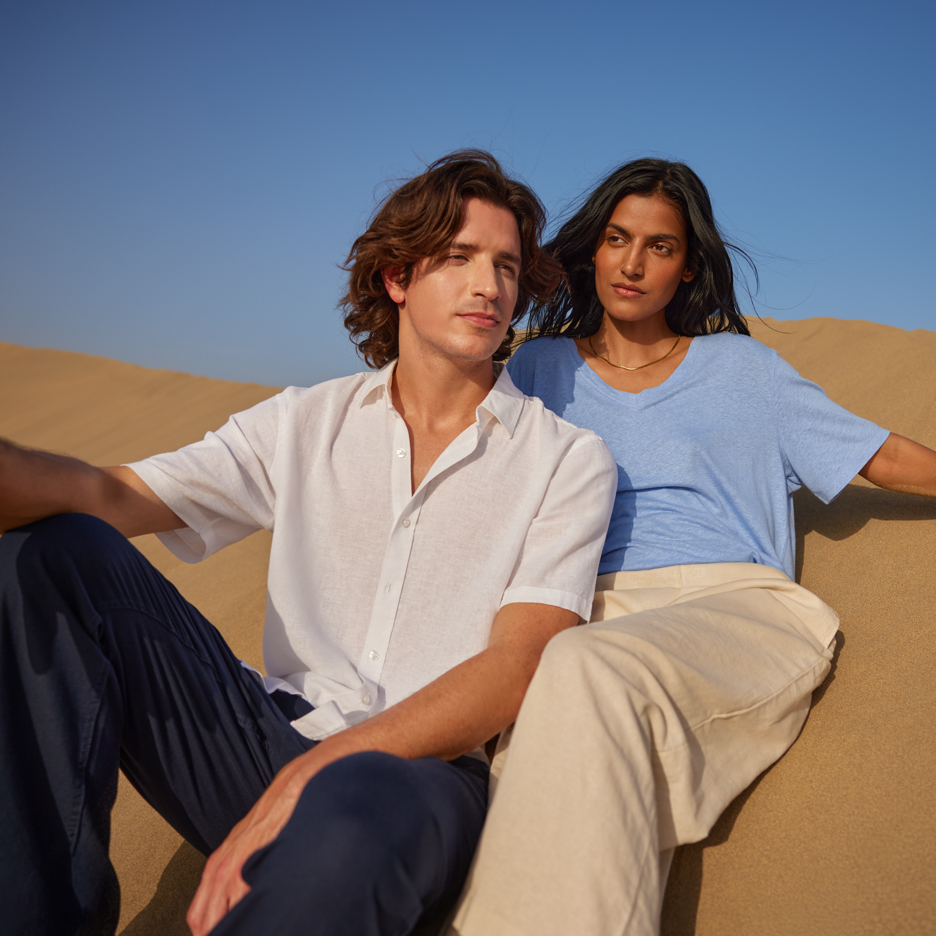 Duas pessoas jovens no deserto, vestindo camisa branca e t-shirt azul, calças azul-marinho e bege.