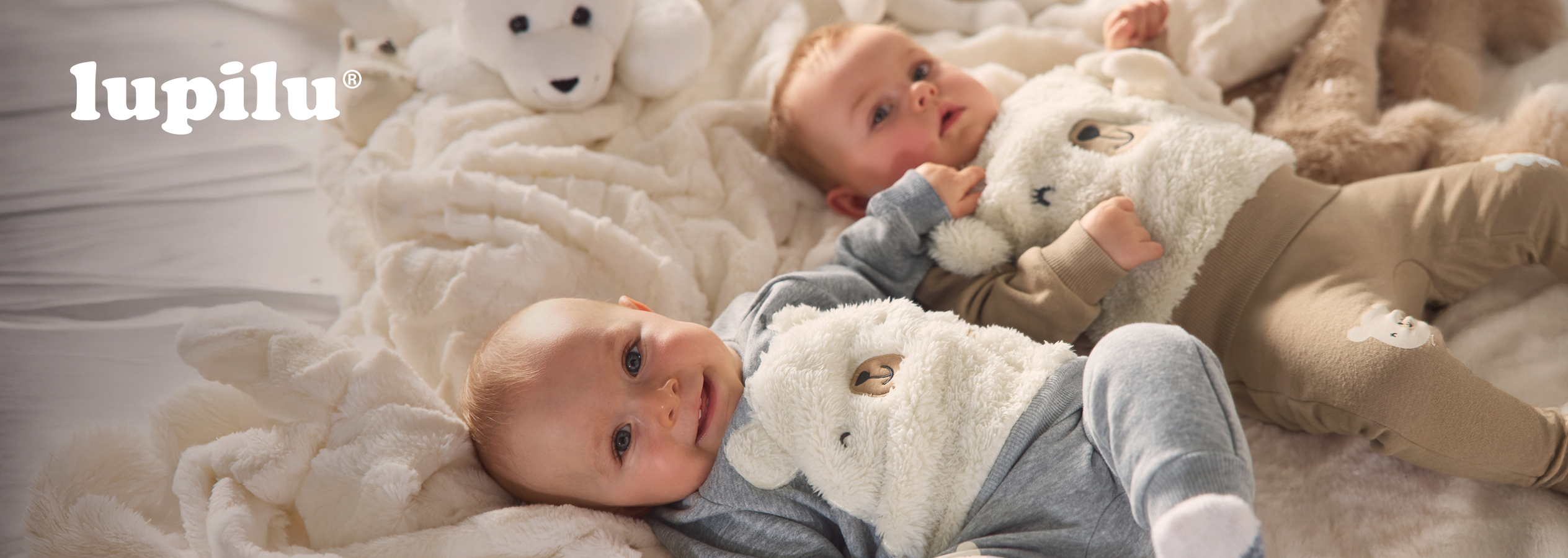 Dois bebés Lupilu deitados numa manta fofa, um com um fato de urso de peluche.