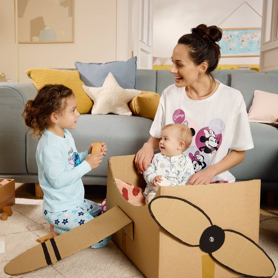 Mãe e filhos brincando com uma caixa de papelão, com a mãe usando uma camiseta de personagens de desenho animado.