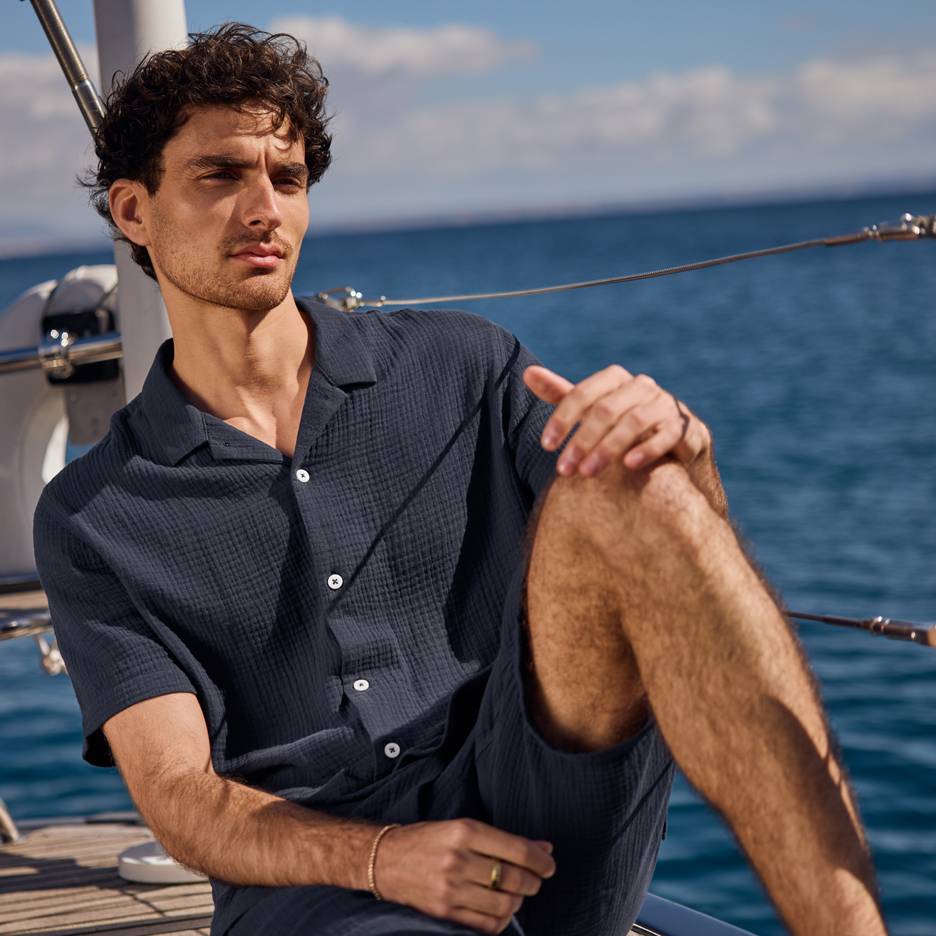 Homem de camisa e calções azul-escuros num iate, a olhar para o mar.