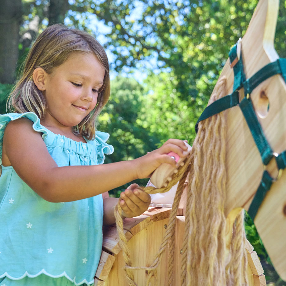 Menina a escovar a crina de um cavalo de baloiço de madeira ao ar livre.