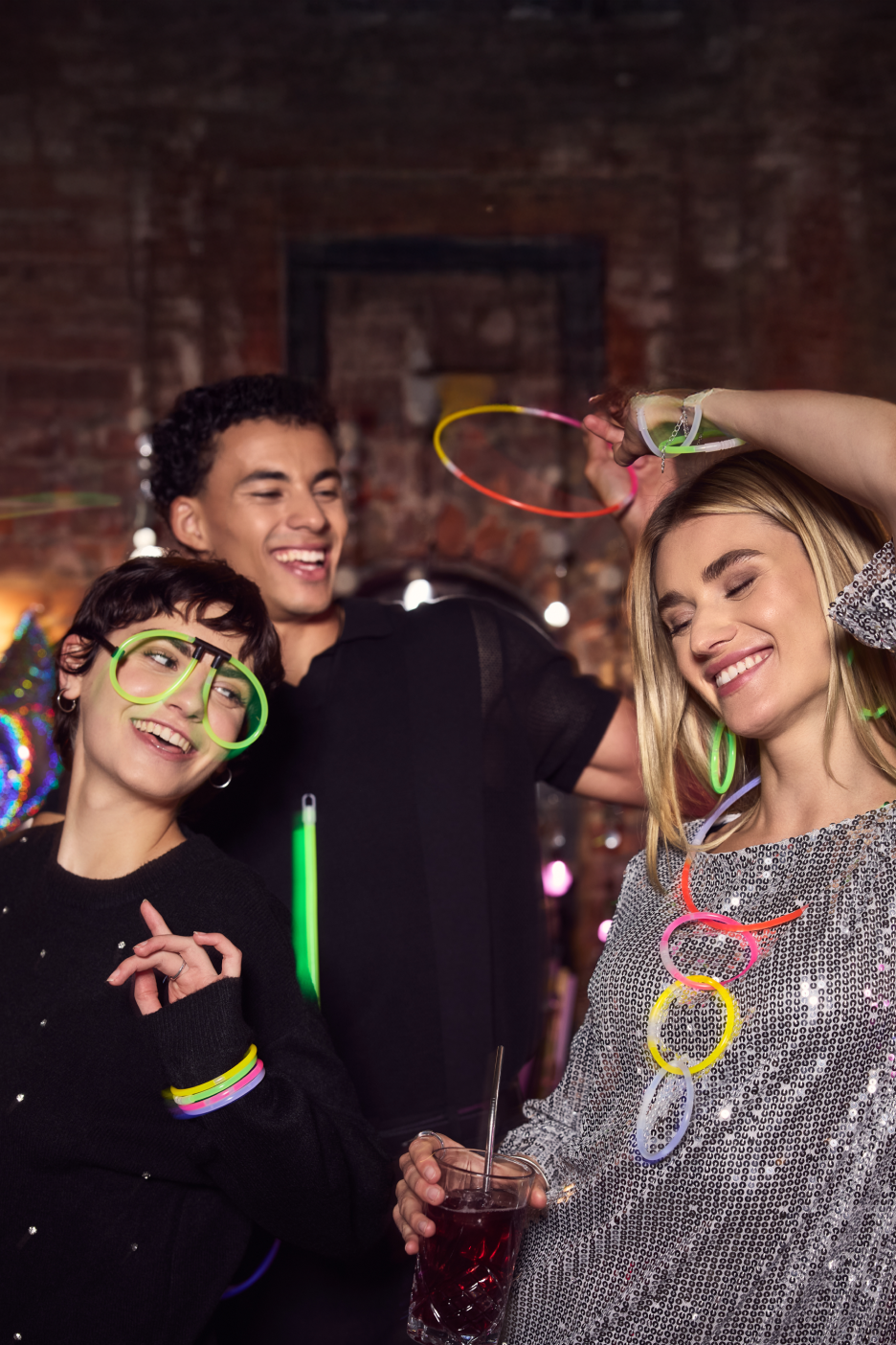 Três amigos a divertirem-se numa festa com óculos e joias que brilham no escuro.