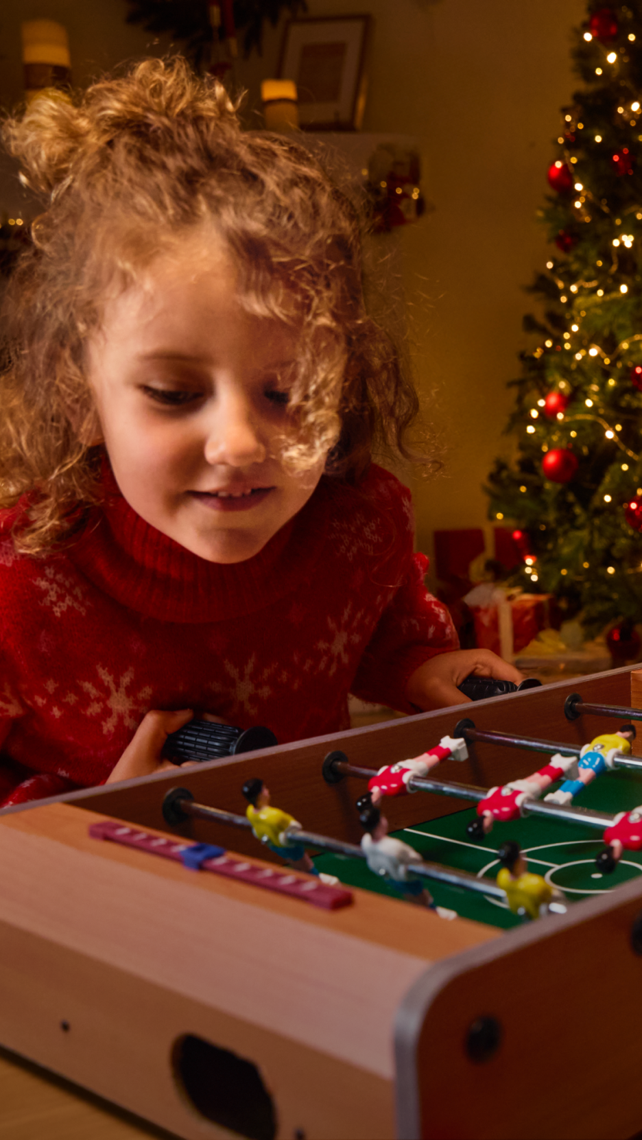 Criança a jogar matraquilhos com uma árvore de Natal iluminada ao fundo.