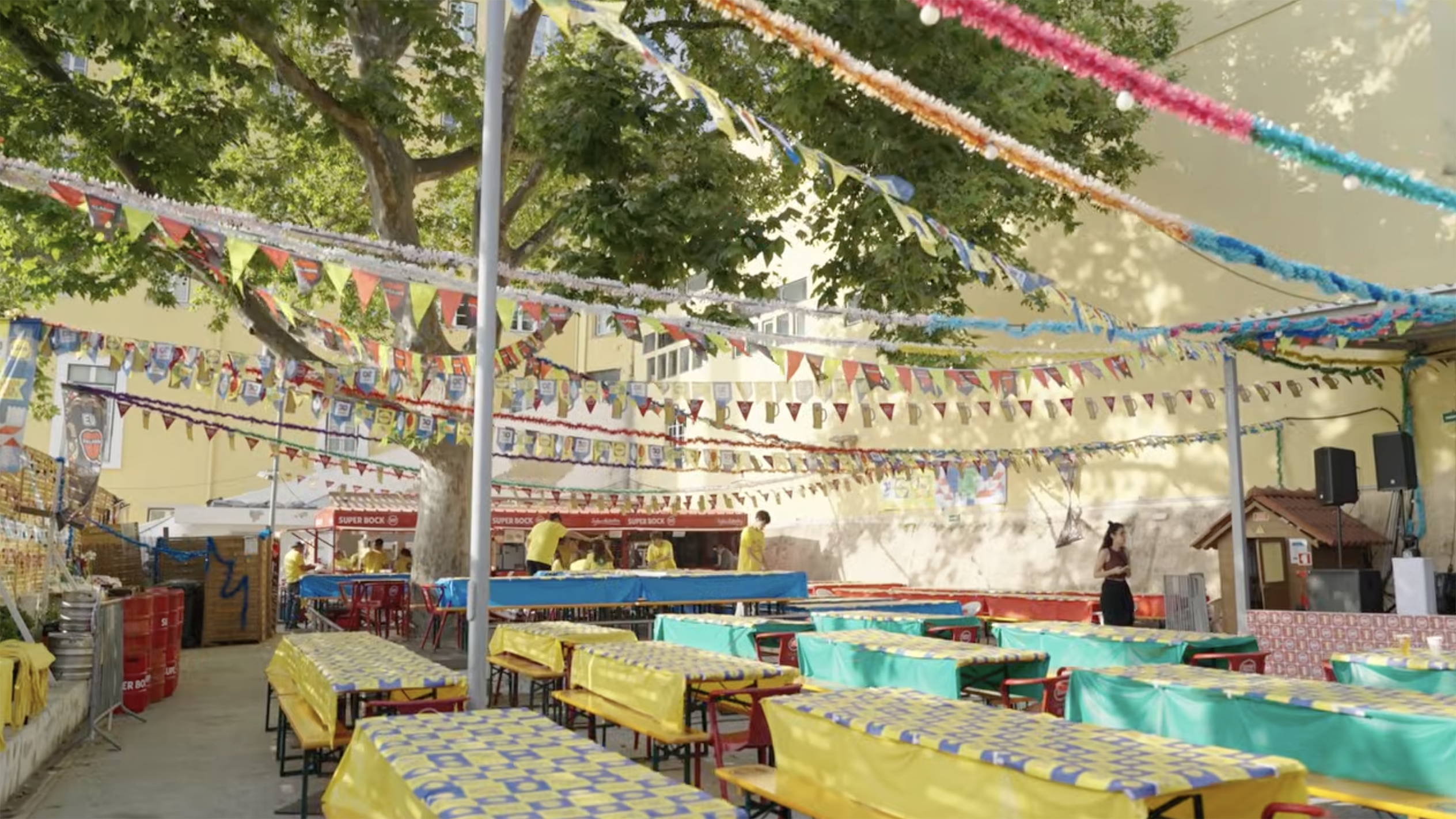 Bancadas de festa com toalhas amarelas e azuis, decoradas com grinaldas e bandeiras.