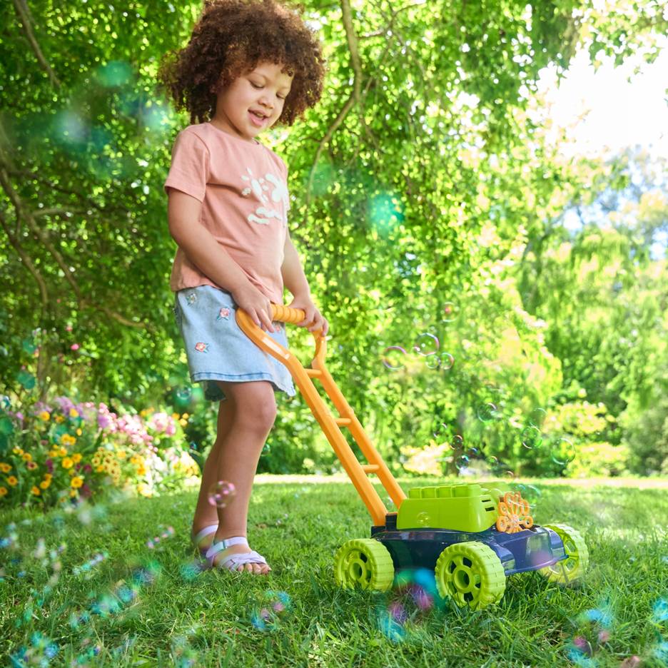 Uma criança com cabelo encaracolado empurra um cortador de grama de brinquedo que faz bolhas na grama verde.