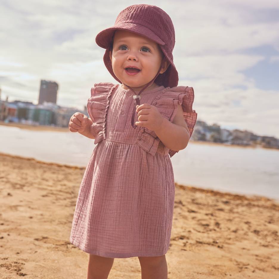 Bebé com vestido rosa e chapéu na praia.