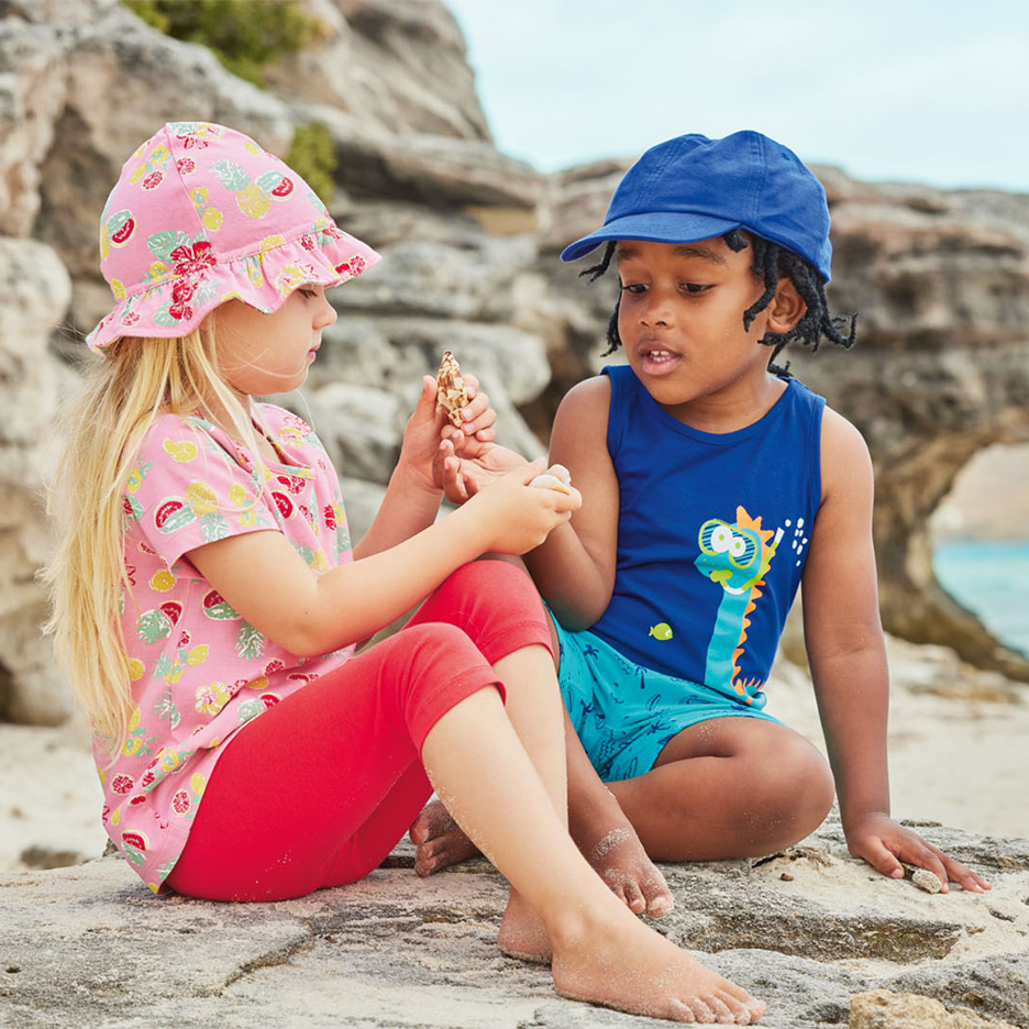 Duas crianças na praia, uma com chapéu de sol rosa e top com padrão de frutas, a outra com boné azul e camisola de dinossauro, segurando conchas.