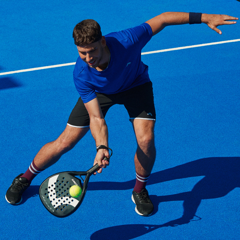 Homem de t-shirt azul e calções pretos a jogar padel com raquete e bola.