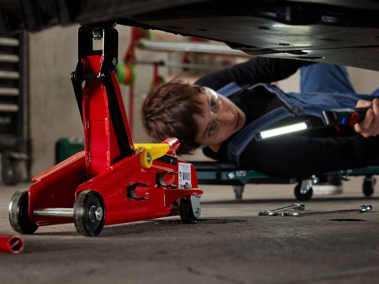 Macaco hidráulico vermelho e mulher debaixo de um carro com luz de trabalho e ferramentas.