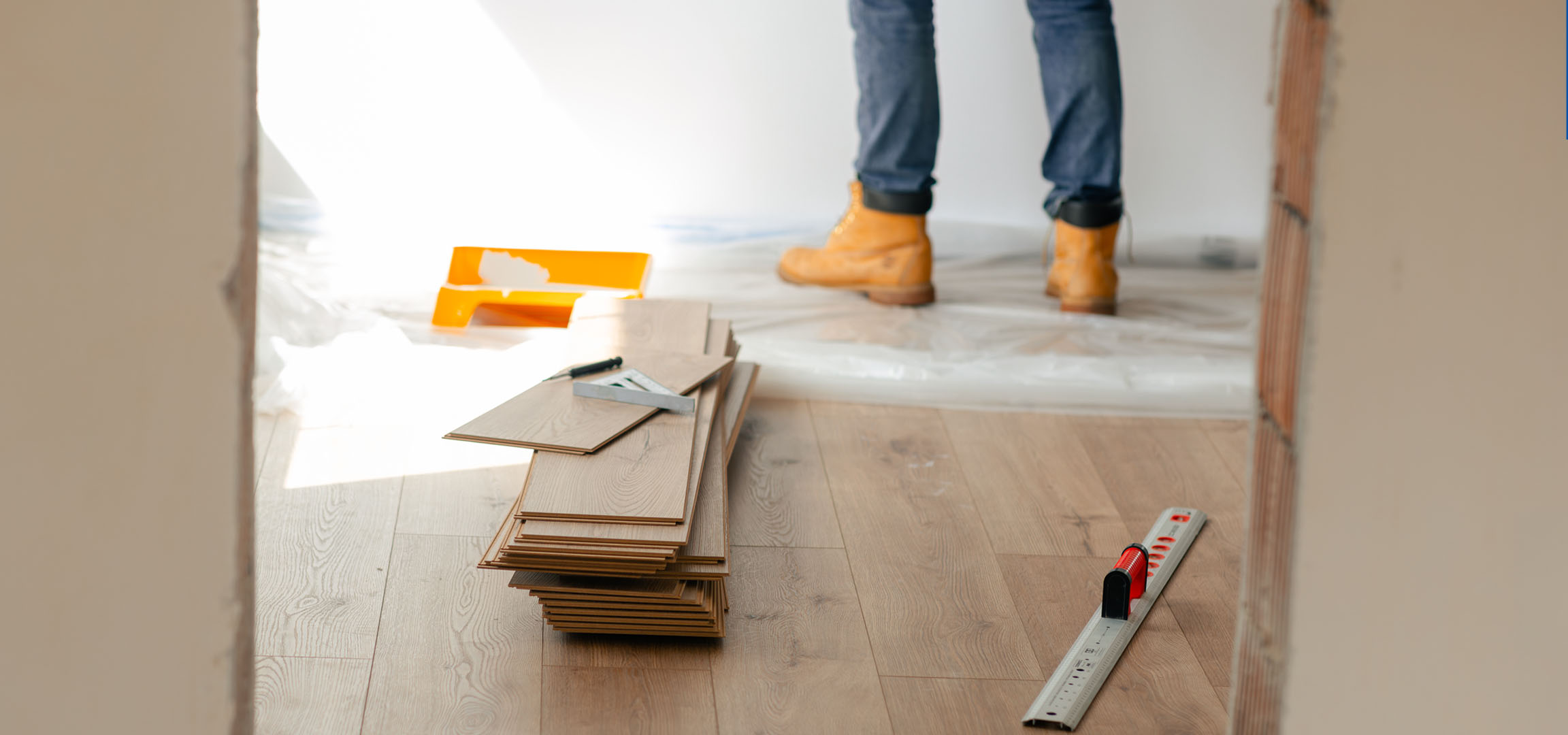 Tábuas de piso laminado e ferramentas de instalação num chão de madeira, com uma pessoa de botas.