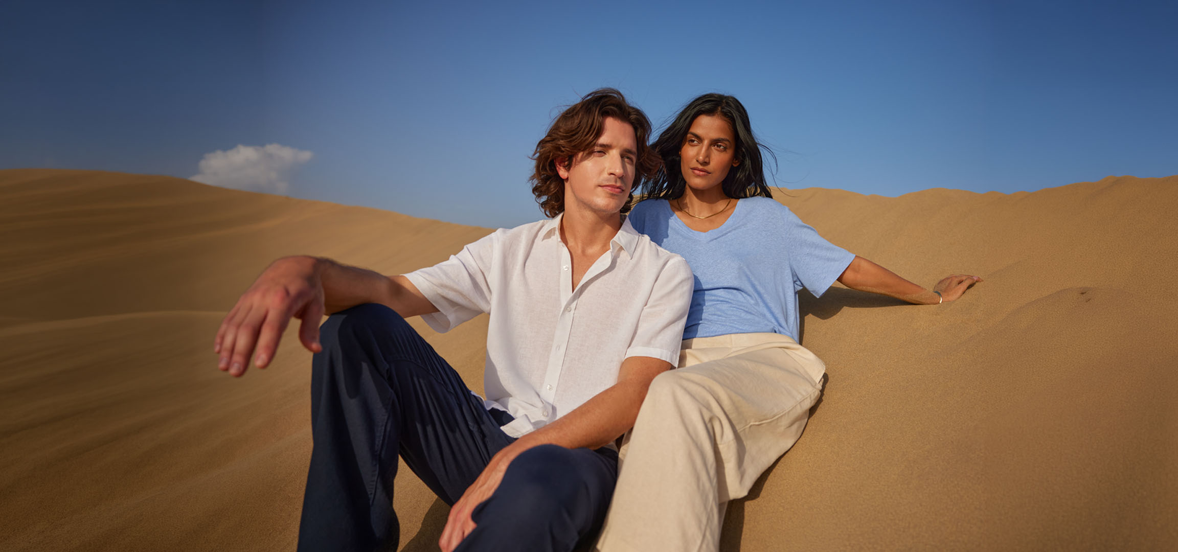 Homem e mulher sentados em dunas de areia, vestindo camisas e calças de linho.