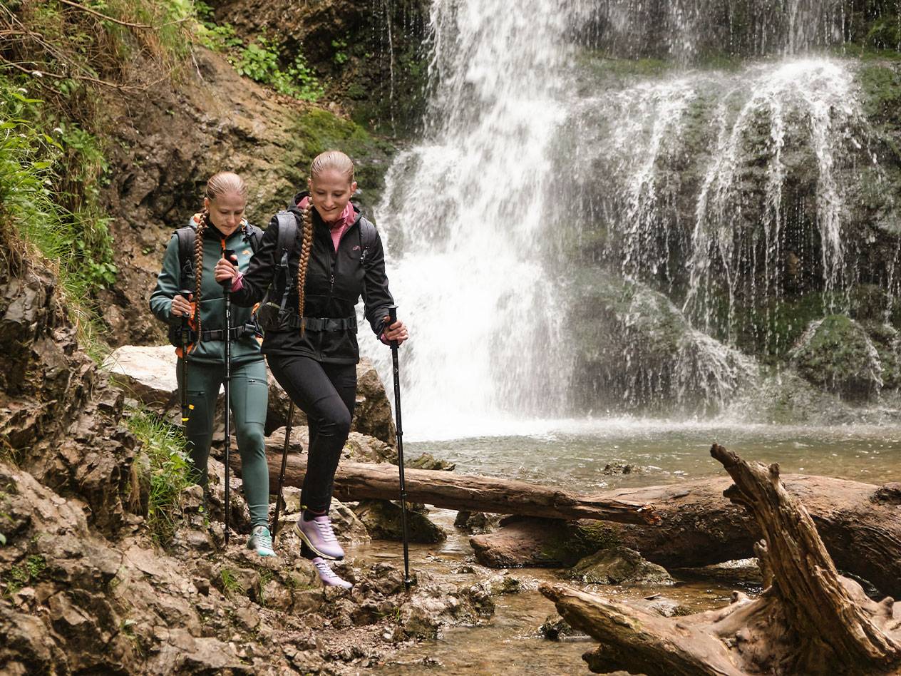 Duas mulheres com bastões de caminhada caminham por um trilho rochoso perto de uma cachoeira.
