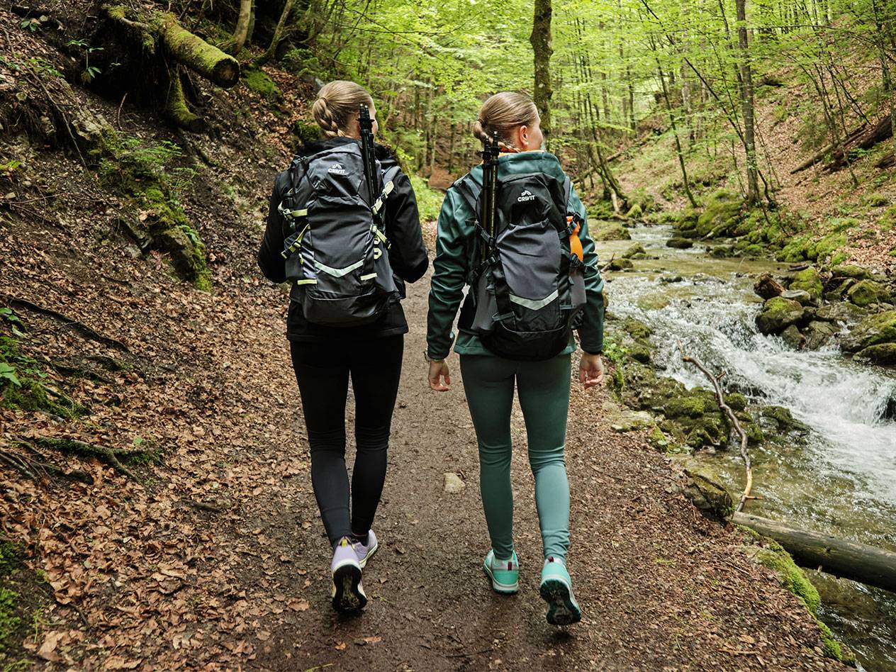 Duas mulheres com mochilas Crivit fazem caminhada por uma trilha na floresta.